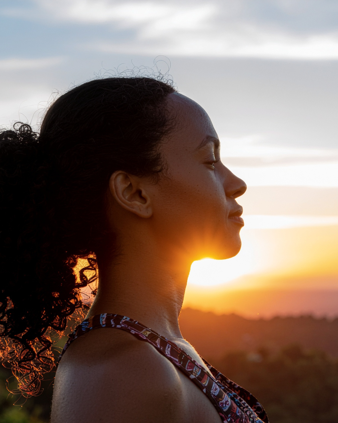 A woman in profile facing a warm sunset, eyes softly closed